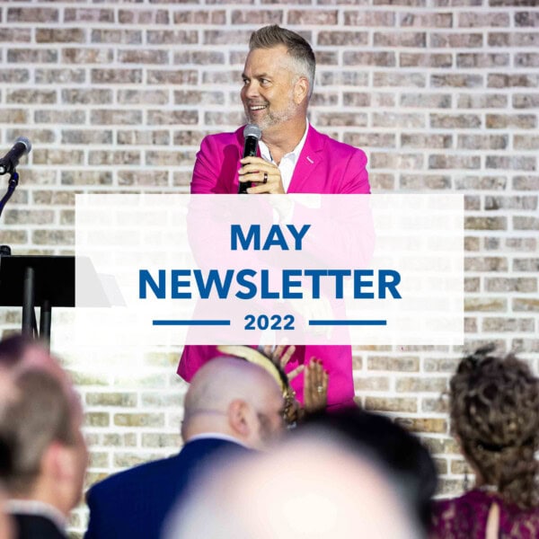 A man in a bright pink blazer speaks into a microphone at an event, standing in front of a brick wall. Overlaid text reads, “May Newsletter 2022.” Audience members are visible in the foreground.