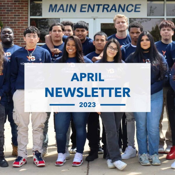A diverse group of teenagers, wearing matching dark blue sweatshirts, stand together and smile in front of a building entrance. A banner in front reads, “APRIL NEWSLETTER 2023.”.