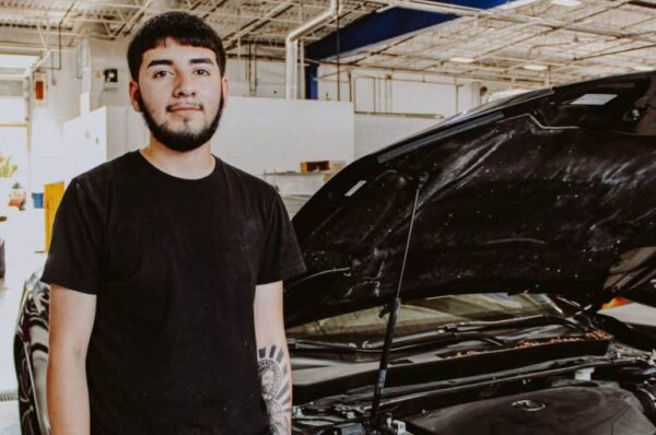 A young man with a beard, wearing a black t-shirt, stands next to a black car with its hood open inside a brightly lit garage or workshop.