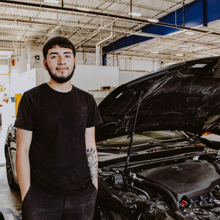 A young man with a beard, wearing a black t-shirt, stands next to a black car with its hood open inside a brightly lit garage or workshop.