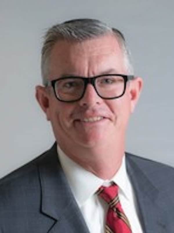 A middle-aged man with short gray hair, wearing black glasses, a gray suit, white shirt, and a red striped tie, smiling at the camera against a plain light background.