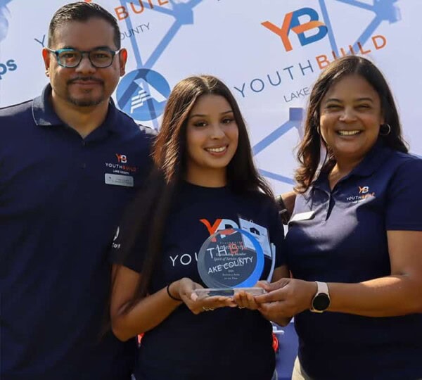 Three people stand smiling in front of a YouthBuild Lake County banner. The young woman in the middle holds a glass award, while the two adults on either side have their arms around her. All wear navy YouthBuild shirts.