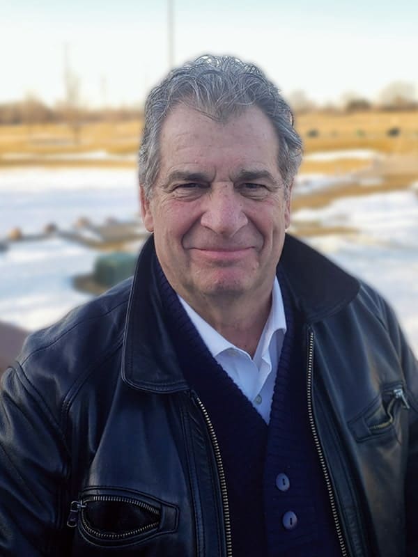 An older man with gray hair smiles outdoors, wearing a black leather jacket over a navy sweater and white collared shirt. Snow and brown grass are visible in the background on a clear day.