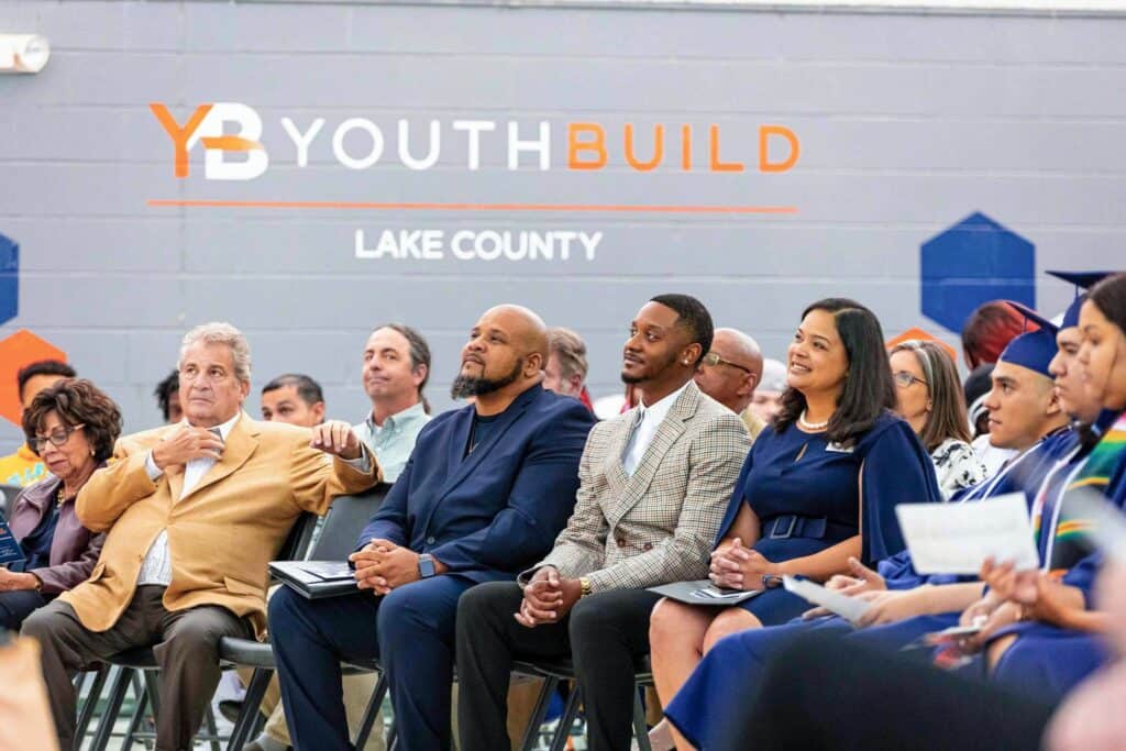 A diverse group of people sits in rows, attentively watching an event at YouthBuild Lake County. The background features a large wall with the YouthBuild logo and geometric designs.