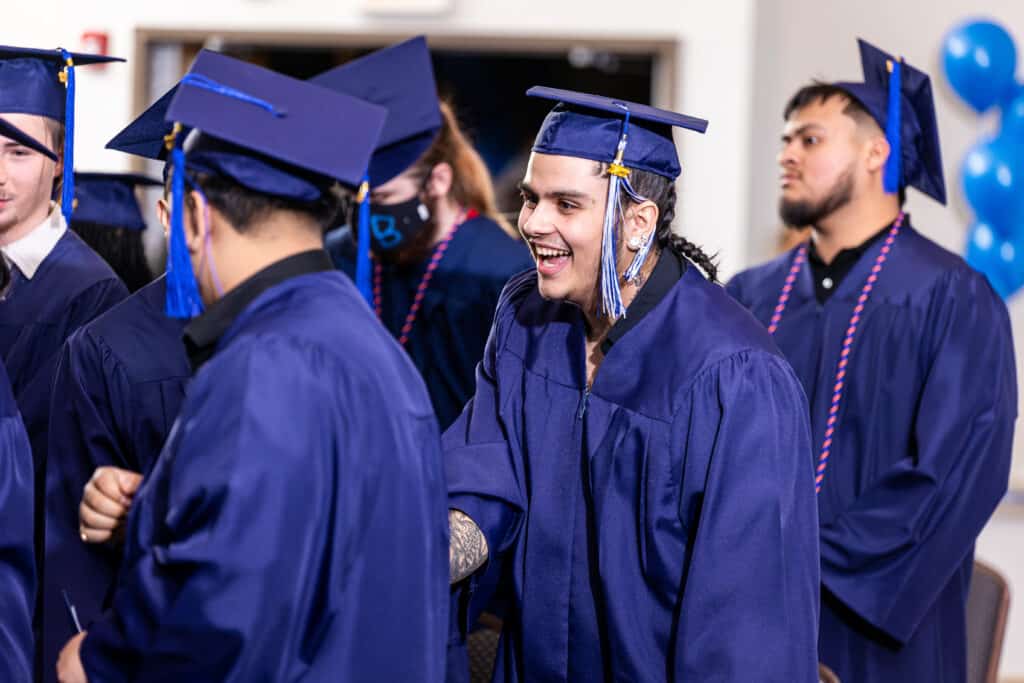 A group of graduates in blue caps and gowns stand together indoors. One smiling graduate is shaking hands with another, while others look on. Blue balloons are visible in the background.