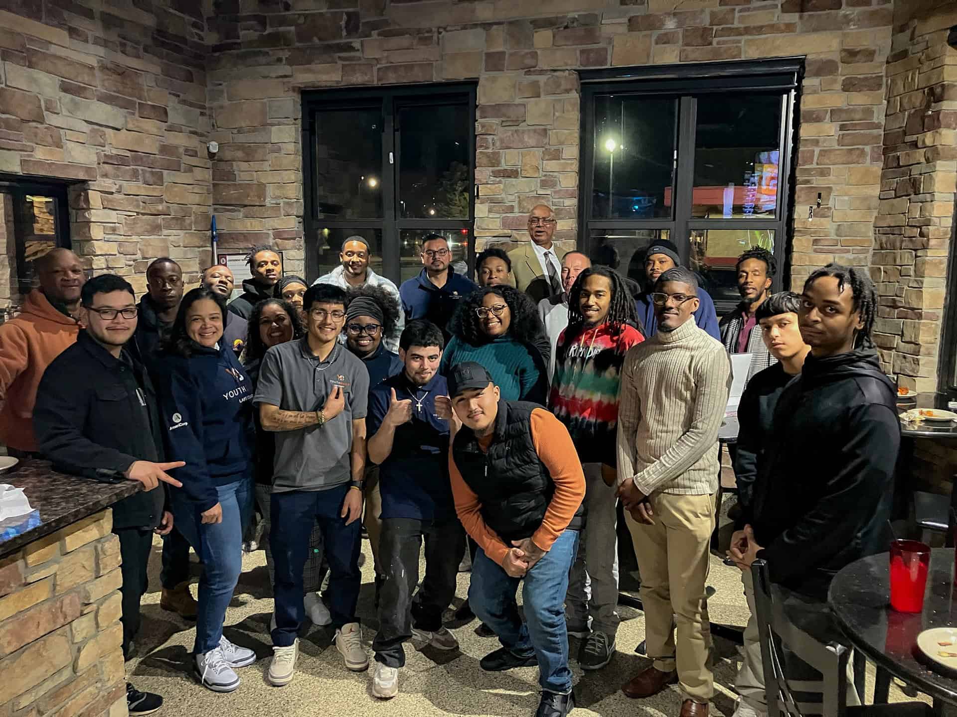 A group of about twenty people, both men and women of diverse ages, are gathered indoors in a warmly lit room with brick walls. They are smiling and posing for a group photo, some making friendly gestures.