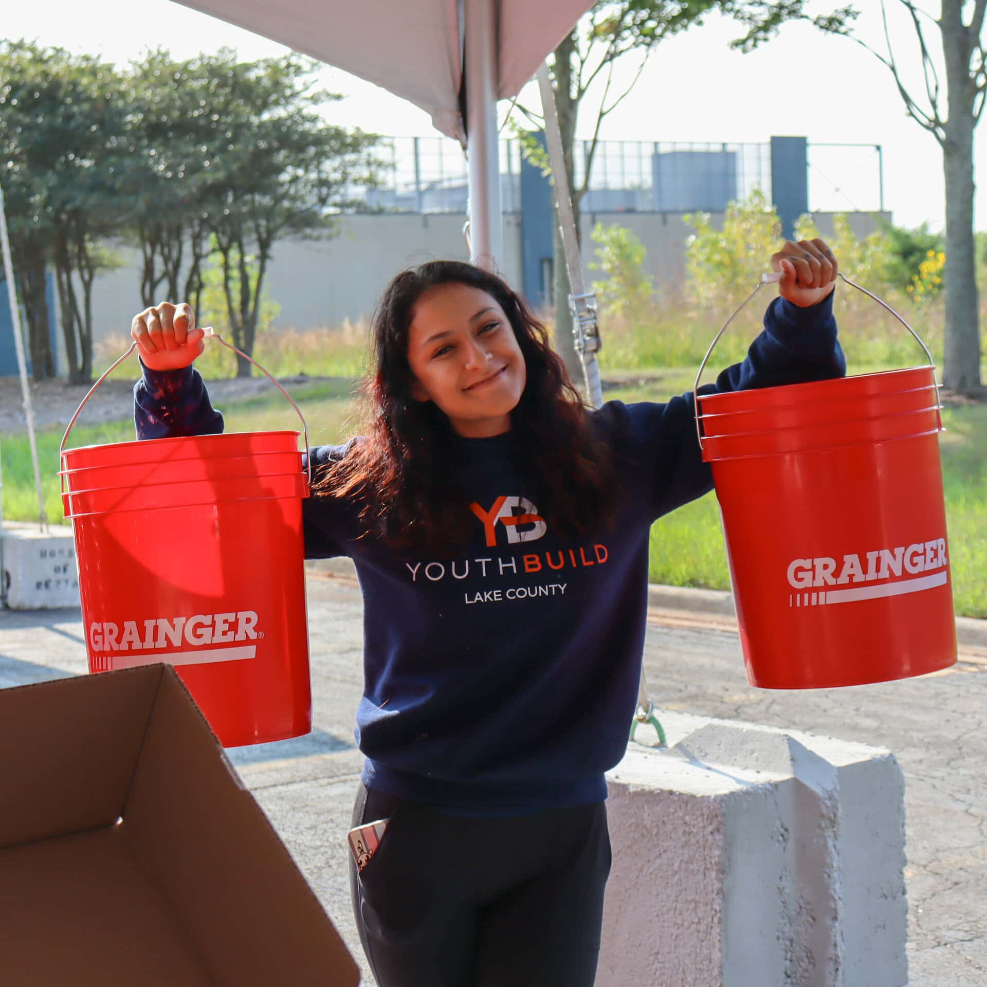 A young woman smiles while holding up two large red buckets labeled Grainger. She is wearing a YouthBuild Lake County sweatshirt and standing outdoors near a tent.