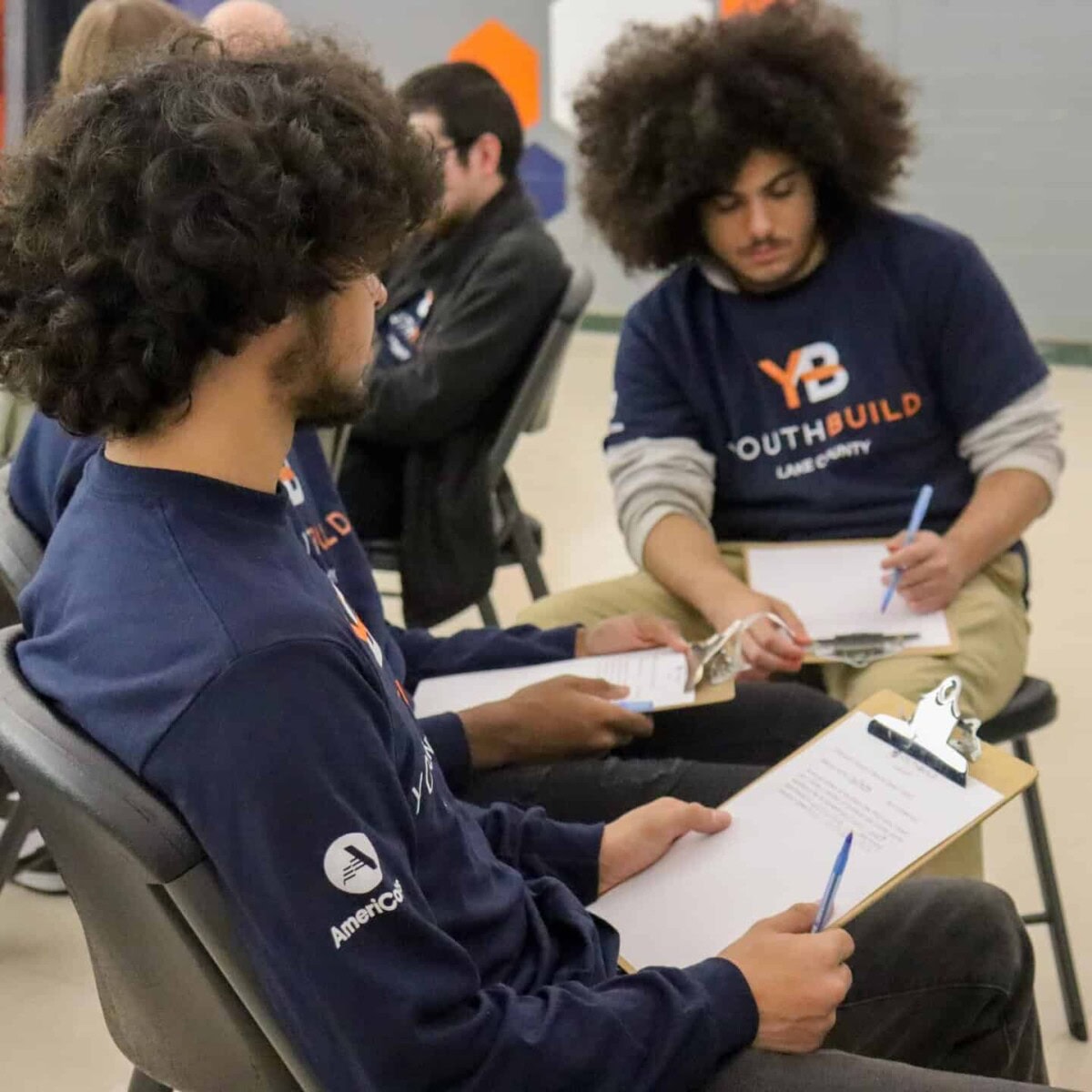 Several young adults sit in chairs facing each other, holding clipboards and writing. They wear matching YouthBuild shirts and appear engaged in a group activity or discussion indoors.