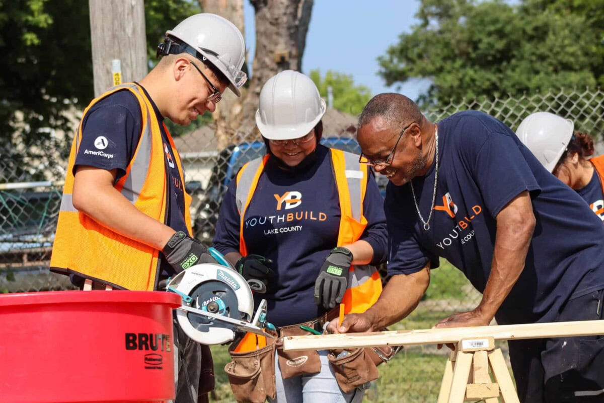 Three people in hard hats and YouthBuild shirts work together outdoors on a construction project, using tools and wood, with a fence and trees in the background.