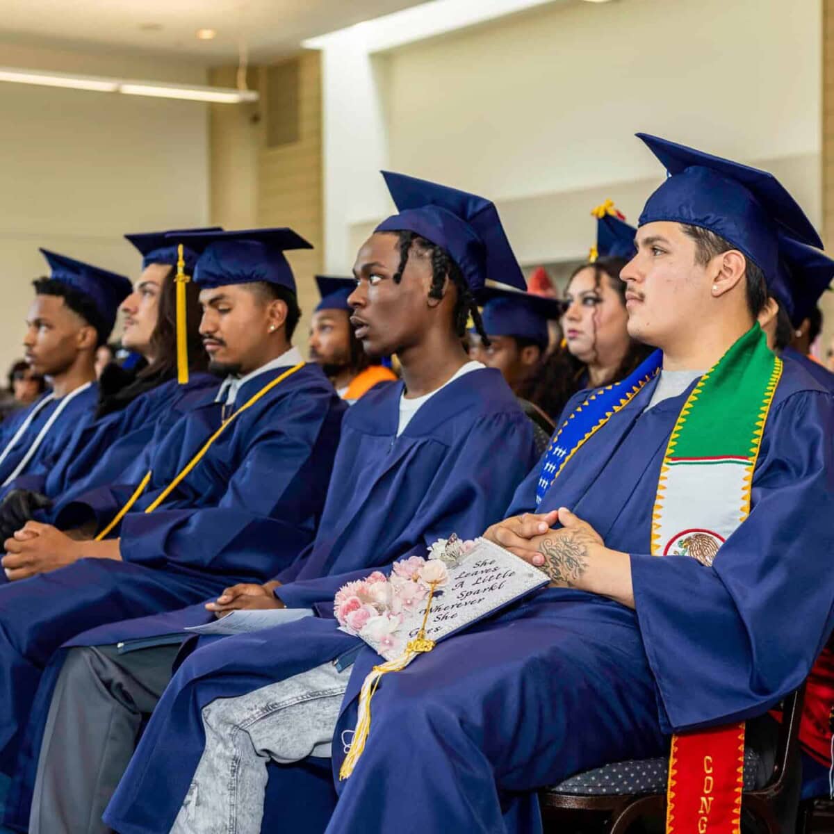 A group of graduates in blue caps and gowns sit in rows at a ceremony, looking forward attentively. One graduate holds flowers, and another wears a colorful sash and stole over their gown.
