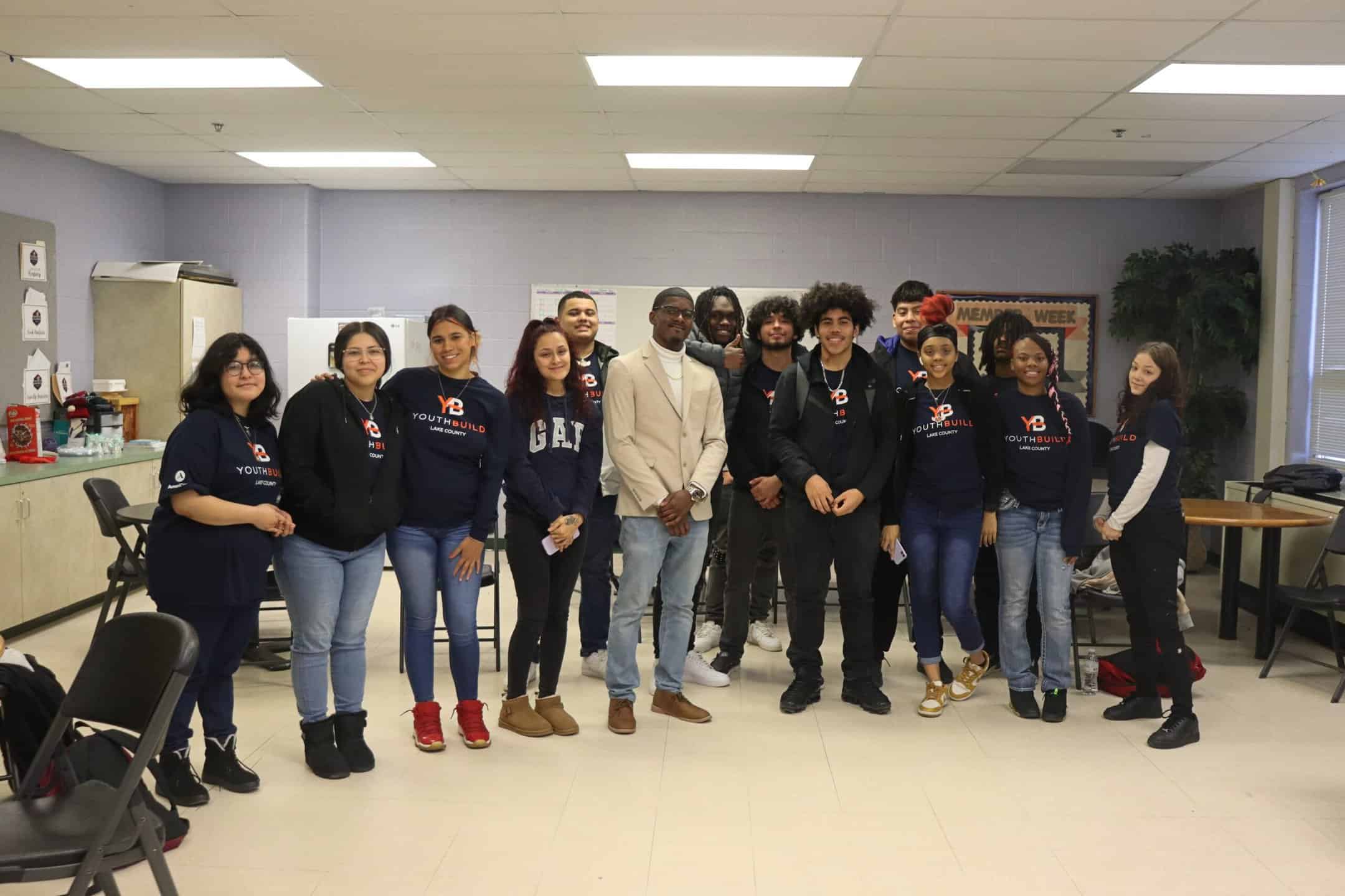 A group of people, mostly young adults, stand together and smile for a photo in a classroom. Many are wearing matching navy “YouthBuild” shirts, and some wear casual clothing. Tables and chairs are visible in the background.