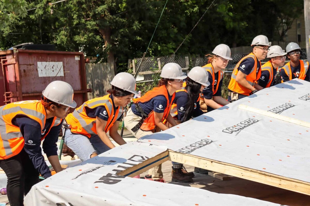 A group of people wearing hard hats and orange safety vests work together to lift and position a large sheet of building material at a construction site outdoors.