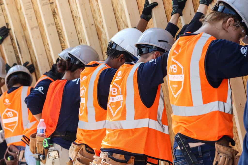 Five construction workers wearing orange safety vests and hard hats lean against a wooden wall, working together on a construction site. They have tool belts and appear to be focused on their tasks.