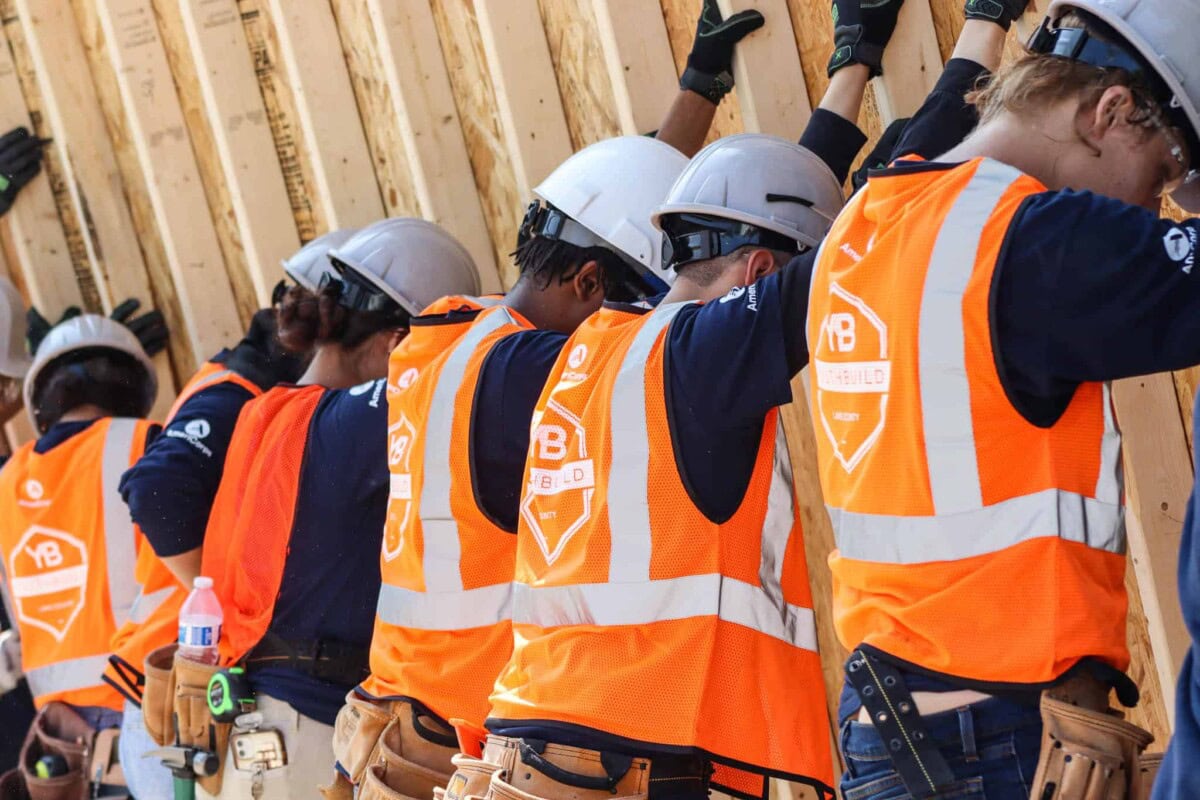 A group of construction workers wearing hard hats and orange safety vests press wooden beams against a wall as part of a building project. Their backs are visible, and they appear to be focused and working together.