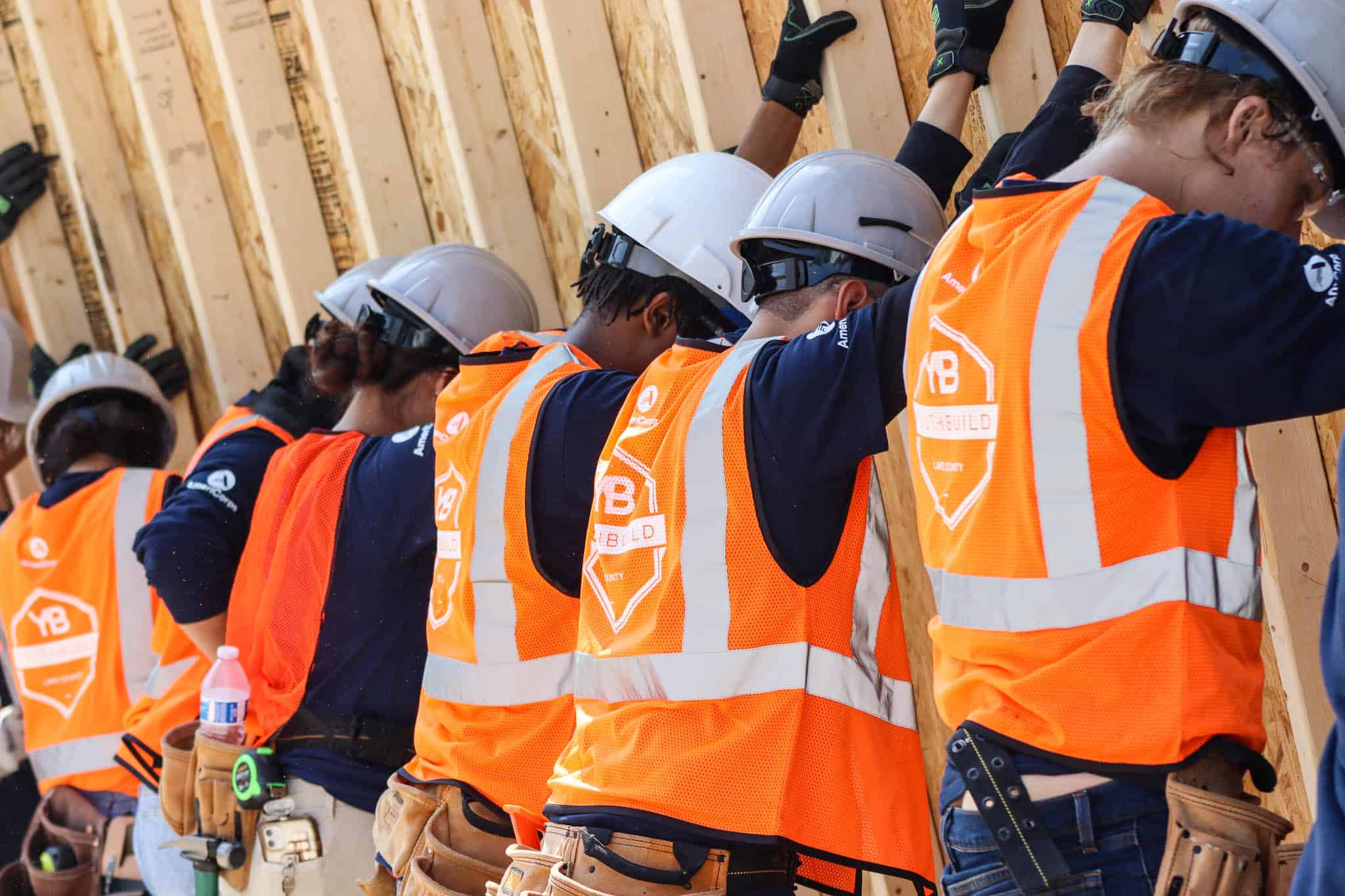 A group of construction workers wearing hard hats and orange safety vests press wooden beams against a wall as part of a building project. Their backs are visible, and they appear to be focused and working together.