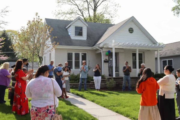 A group of people stand on a lawn in front of a small white house, some clapping and some holding phones, during a sunny outdoor gathering. Balloons are tied to the front porch.