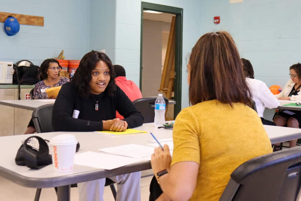 Two women sit across from each other at a table, engaged in conversation. Papers, a water bottle, and a coffee cup are on the table. Other people sit and talk at tables in the background in a light blue room.