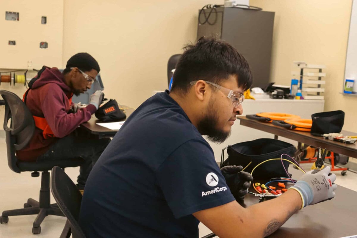 Two young men sit at separate desks in a classroom. The man in front is working with electronics tools and wires, wearing safety glasses and an AmeriCorps shirt. The man in back writes on paper, focused on his task.