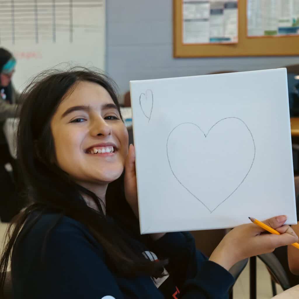 A smiling girl holds up a white canvas with a large heart and a smaller heart sketched on it in pencil, seated at a table with a pencil in hand in a classroom setting.