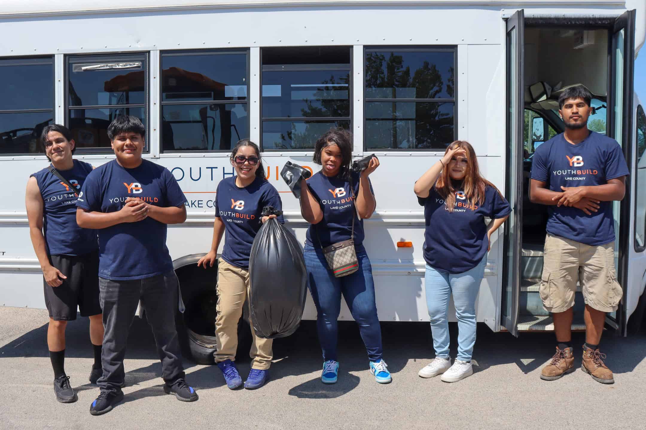 Six young people wearing matching YouthBuild shirts stand smiling in front of a white bus, holding trash bags and cleaning supplies, appearing to participate in a community service project.