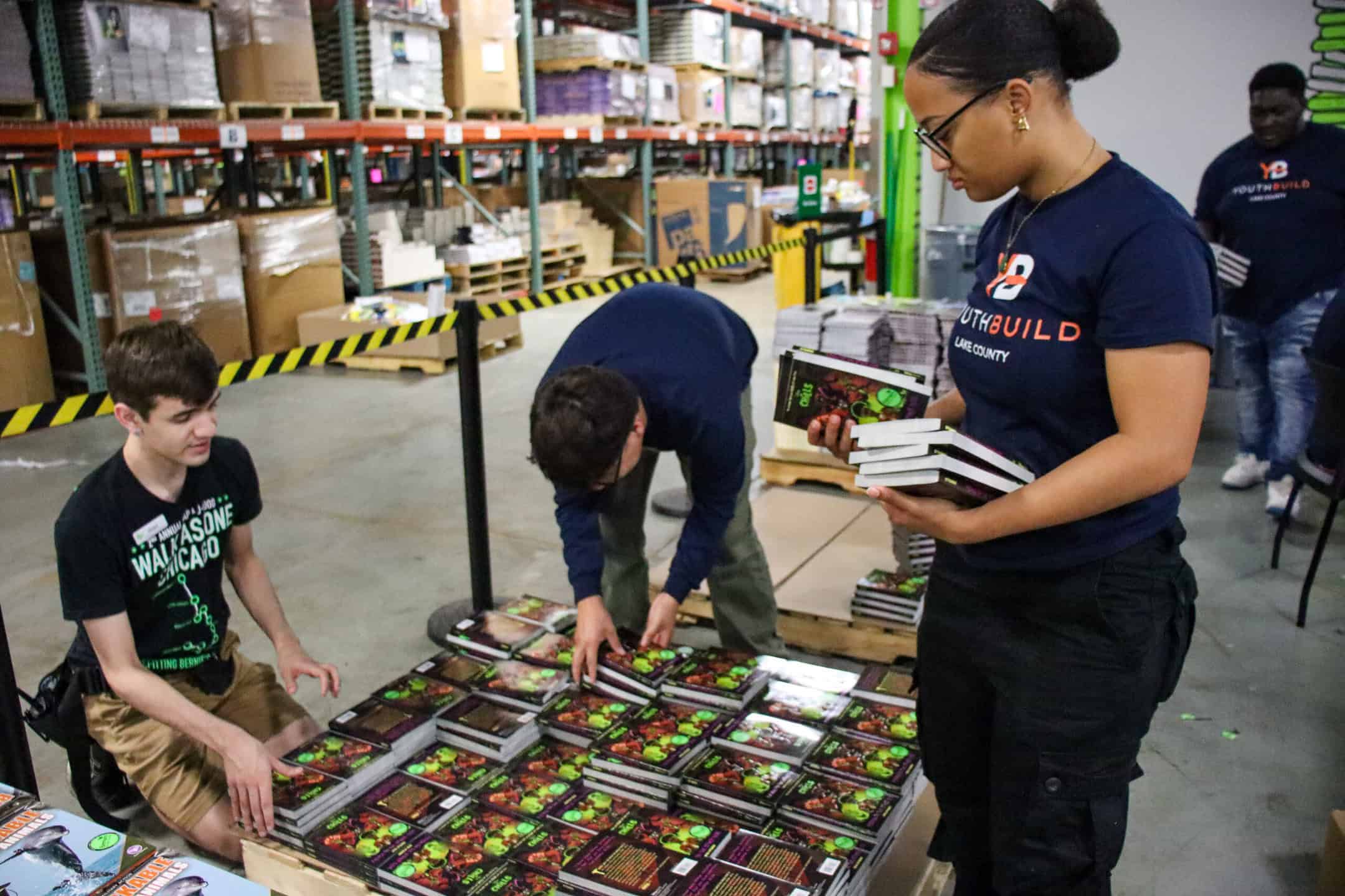 Three young people sort and stack hardcover books on pallets in a warehouse. One person stands holding several books while two others organize books on the table. Shelves filled with boxes are visible in the background.