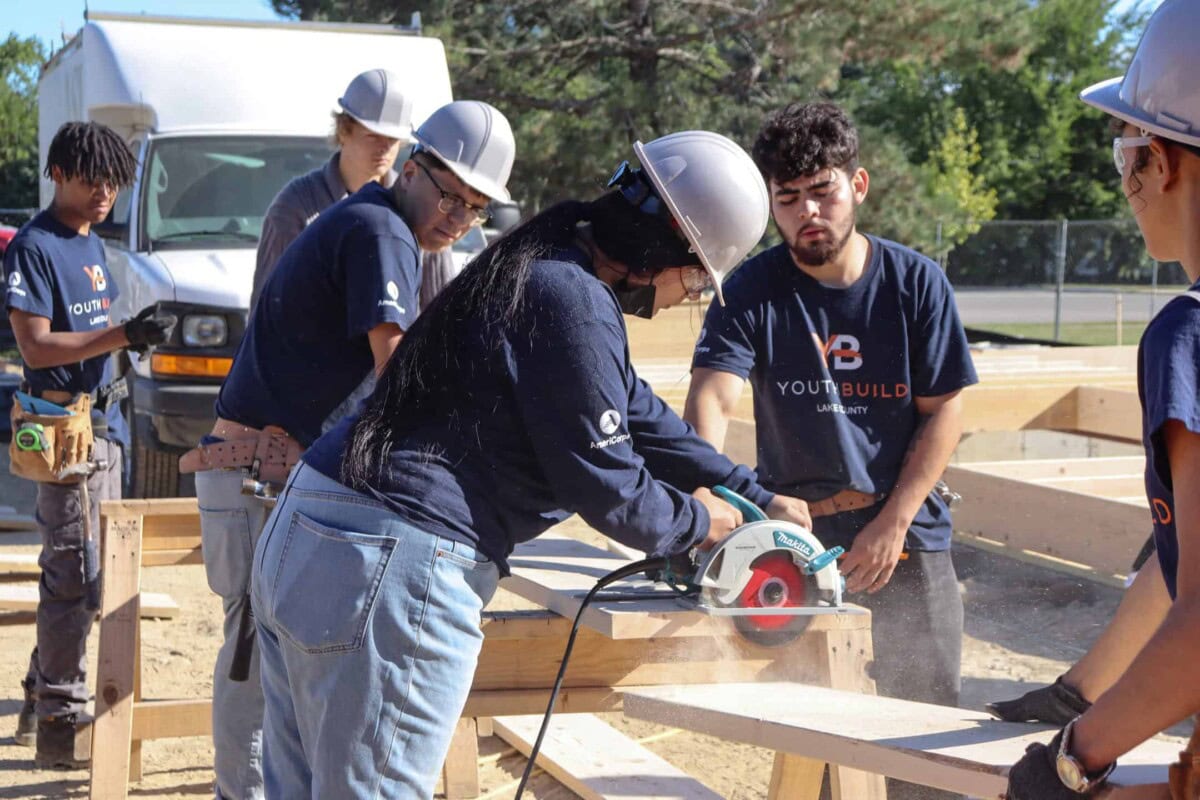 A group of young people in hard hats and YouthBuild shirts work together on a construction project outdoors, with one person using a circular saw to cut wood while others observe and assist.