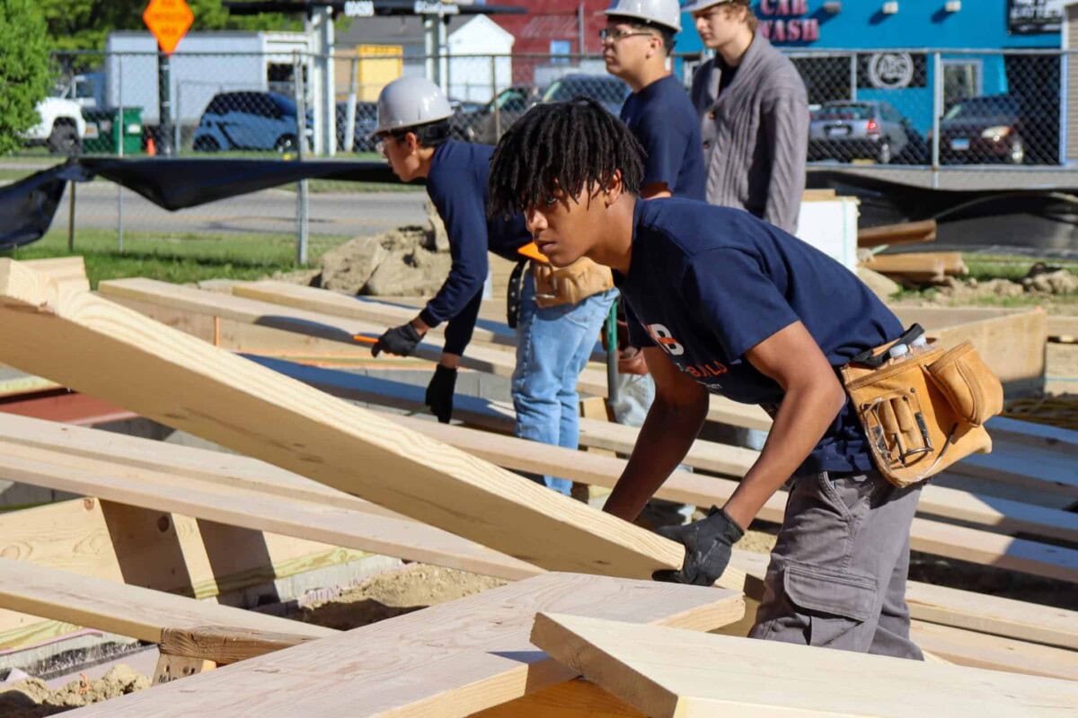 Four construction workers wearing safety gear and tool belts are working with wooden beams at an outdoor construction site, assembling a structure on a sunny day.