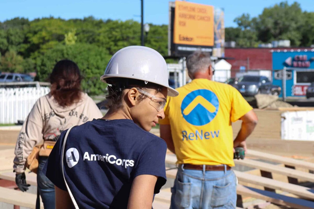A person wearing a white hard hat, safety glasses, and an AmeriCorps shirt stands at a construction site with others, surrounded by wooden beams and outdoor scenery.