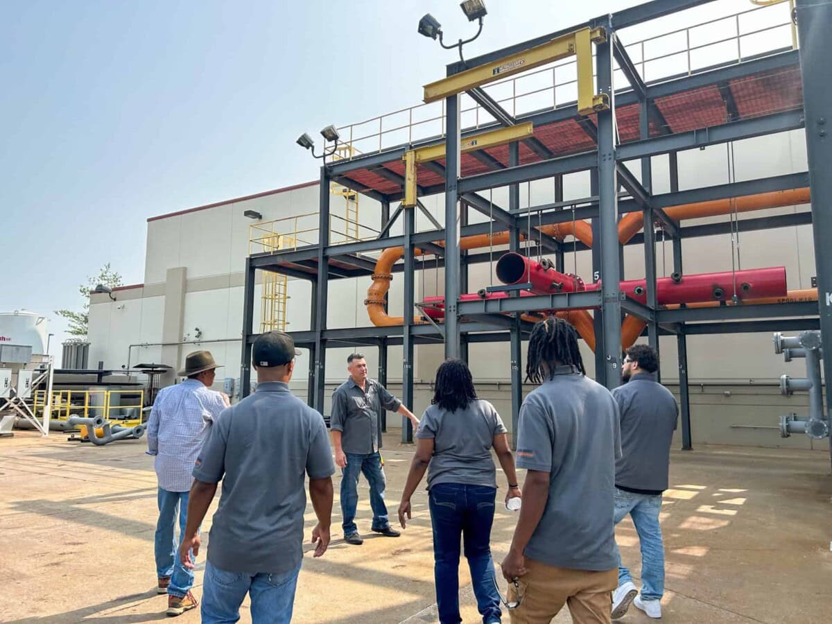 A group of six people in gray shirts observe a man giving a tour outside an industrial facility with metal structures, orange and red pipes, and an overhead walkway. The scene appears to be a workplace or training session.