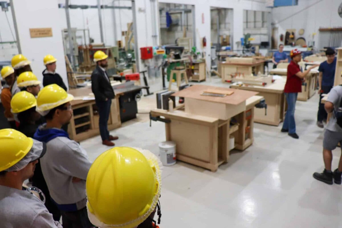 A group of people wearing yellow hard hats listens to an instructor in a workshop filled with woodworking equipment and tables. The instructor stands near the center, gesturing while speaking.