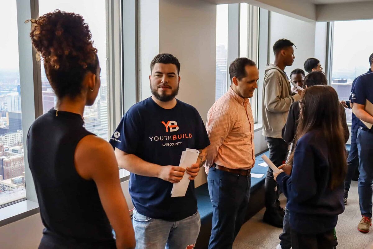 A group of people participates in a networking event in a high-rise office with large windows overlooking a city. They are talking in small groups, some holding papers, and appear engaged and friendly.