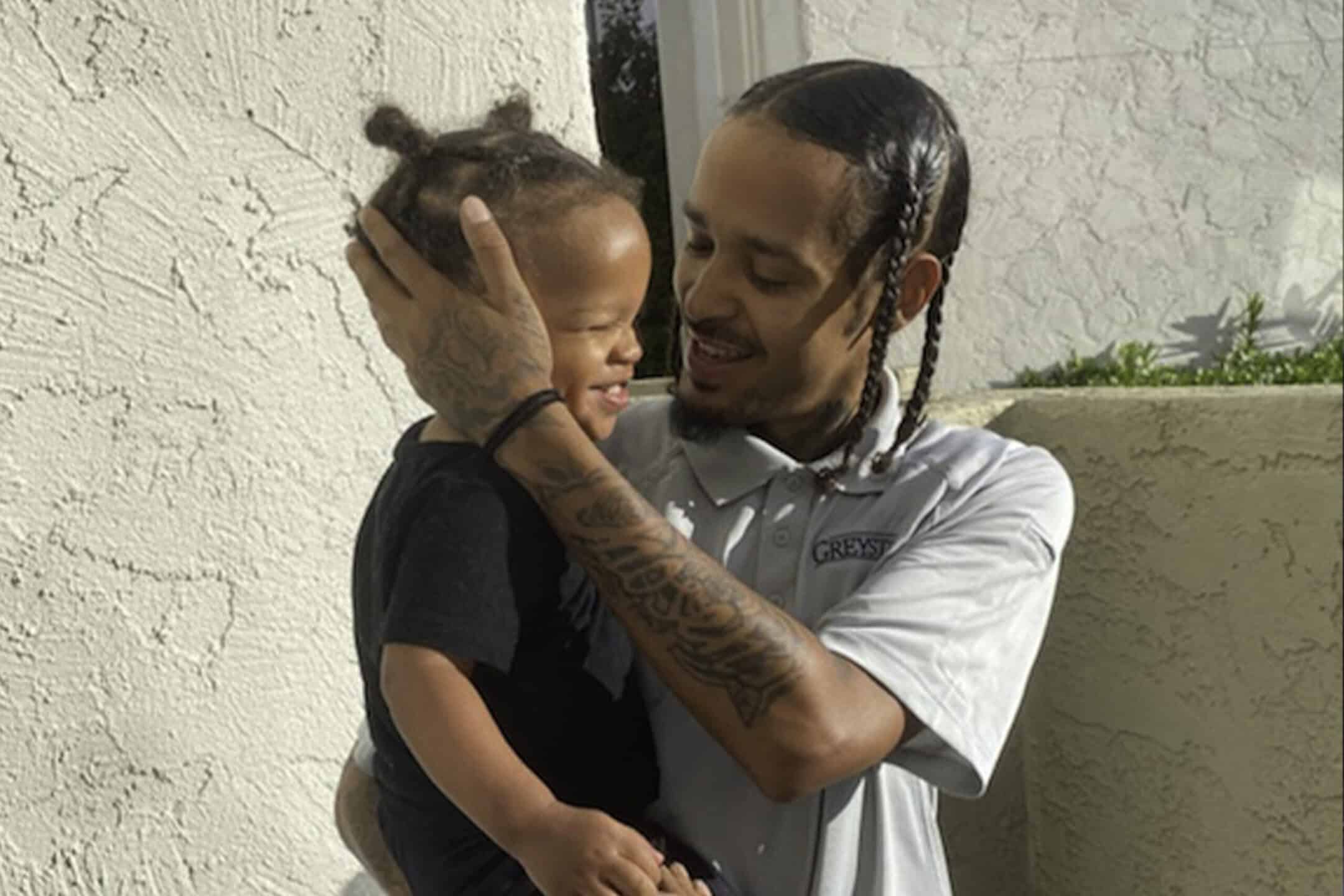 A man with braided hair and tattoos gently holds and smiles at a young child with two small buns, standing together outside near a light-colored wall.
