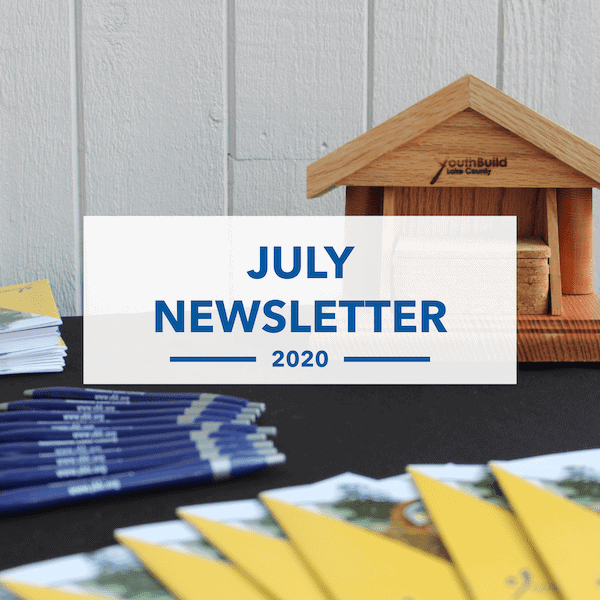 A sign reading July Newsletter 2020 is displayed in front of a small wooden house model. Blue pens and yellow pamphlets are spread out on a black table in the foreground. White wooden wall in the background.