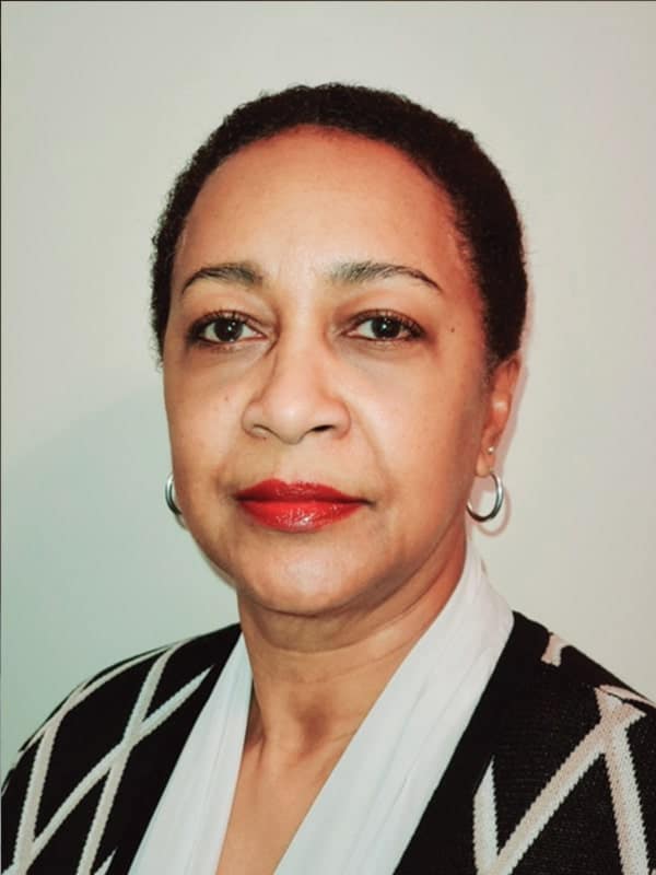 A woman with short hair and hoop earrings, wearing a black and white patterned top over a white blouse, looks at the camera with a neutral expression and red lipstick, against a plain light background.