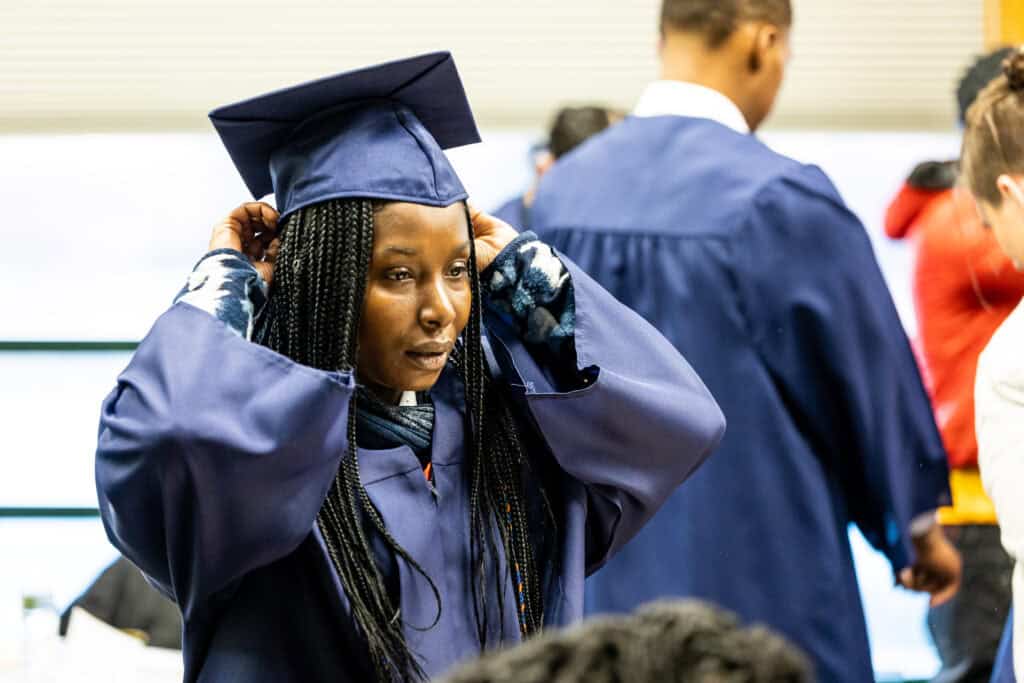 A graduate in a blue cap and gown adjusts her cap, surrounded by other graduates in similar attire at a graduation ceremony.