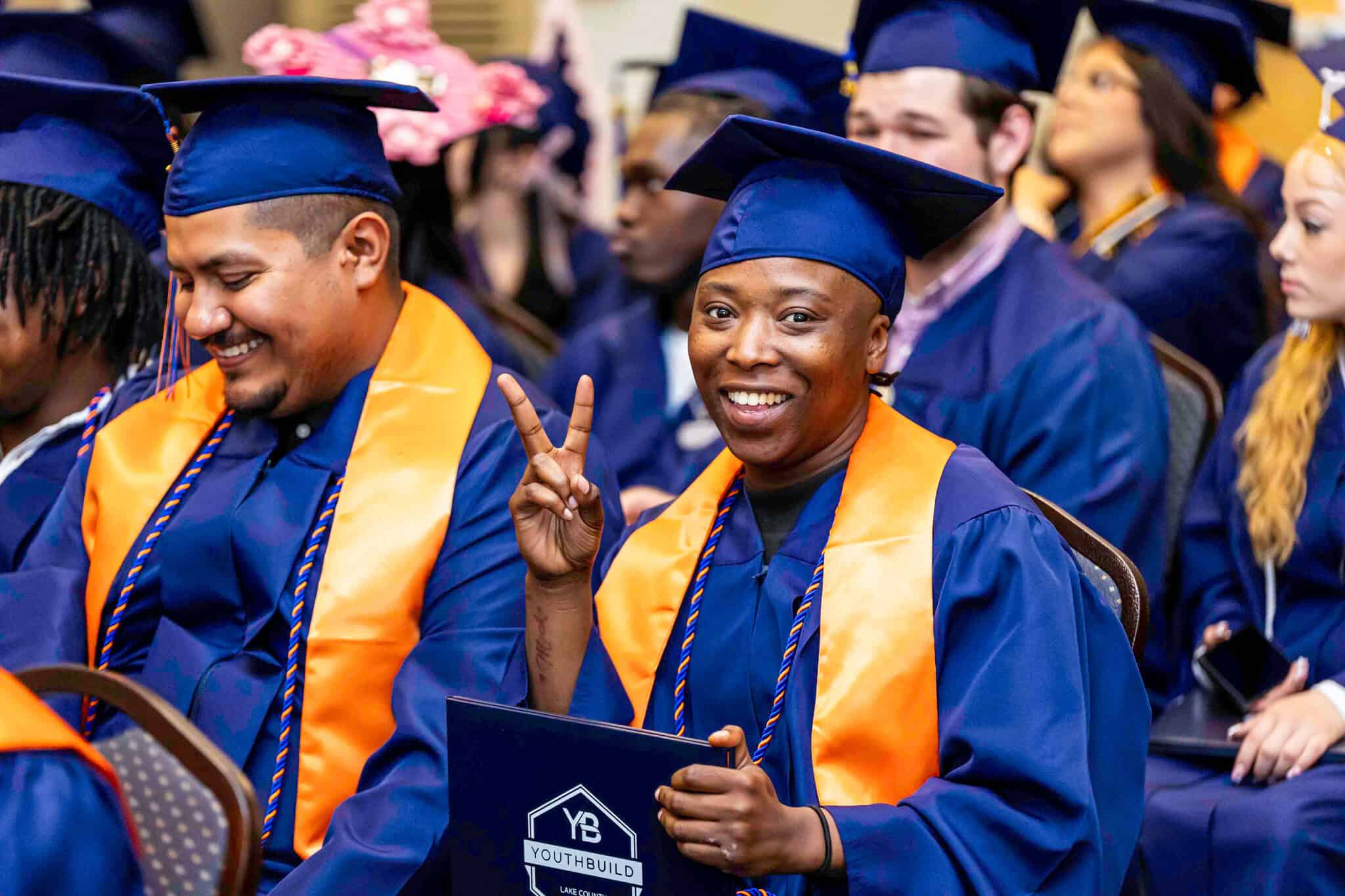 Two graduates in blue caps and gowns with orange stoles sit smiling at a ceremony. One person holds a certificate and makes a peace sign, while the other looks down, also smiling. Other graduates are visible in the background.