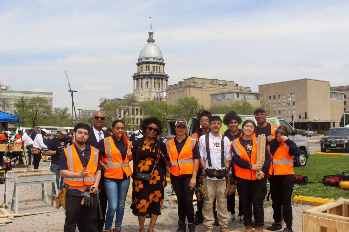 A group of people wearing orange safety vests pose and smile outdoors in front of the Illinois State Capitol building, with tools and construction activity visible in the background.