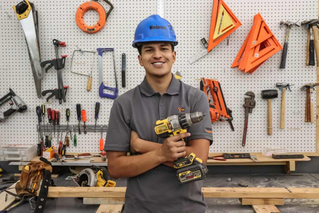 A smiling man in a blue hard hat and gray polo shirt stands in a workshop, holding a cordless drill. Behind him, various tools hang on a pegboard, including saws, squares, and hammers.