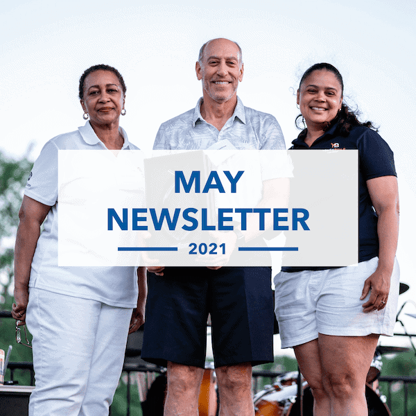 Three smiling adults stand outdoors, two women and one man, behind a transparent banner reading May Newsletter 2021. The group appears happy and casually dressed, with a blue sky in the background.