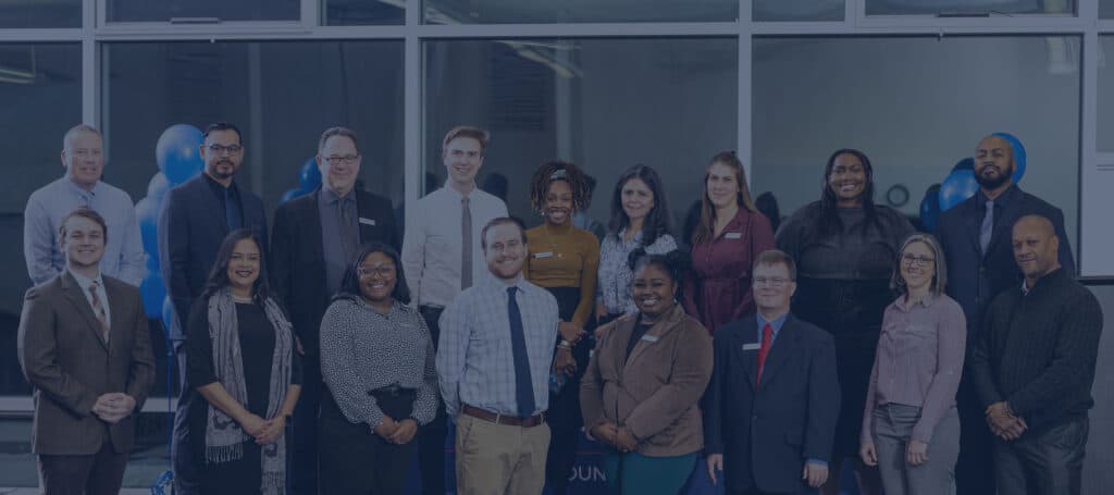 A group of sixteen professionally dressed people poses and smiles for a photo indoors, standing in two rows in front of large windows with blue balloons visible in the background.