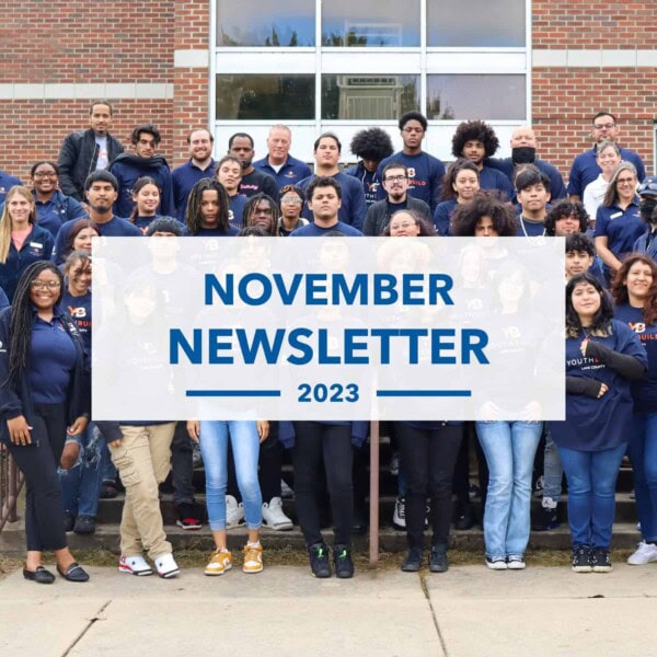 A diverse group of people, mostly young adults and teens, stand together on steps outside a brick building. A large sign in front reads, “November Newsletter 2023.”.