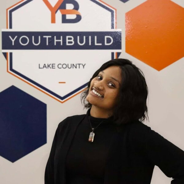 A smiling person with shoulder-length dark hair stands in front of a YouthBuild Lake County sign, wearing a black top and necklace. The background features blue and orange hexagons.