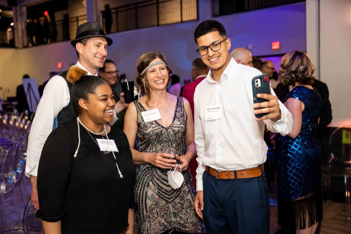 Four people dressed in semi-formal and retro-themed attire smile and pose together for a selfie at an indoor event or party, with other guests visible in the background.