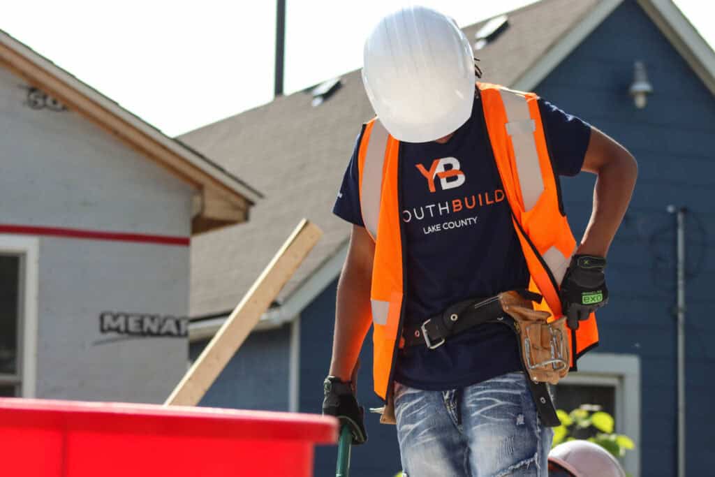 A construction worker wearing a white hard hat, orange safety vest, and tool belt looks down while working outdoors. Their shirt reads YB YouthBuild Lake County. Houses and construction materials are visible in the background.