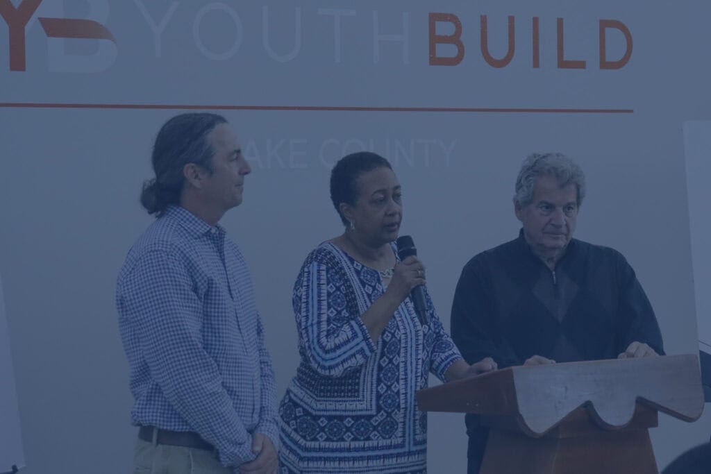 Three adults stand at a podium. A woman in the center holds a microphone and speaks, while two men stand on either side. The background displays the words YOUTH BUILD.