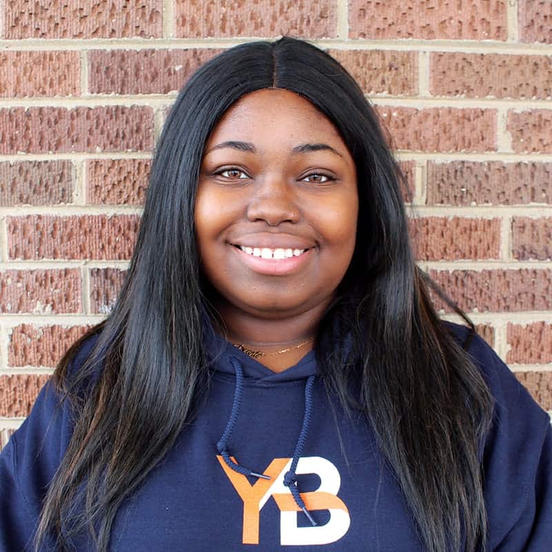 A woman with long, straight black hair smiles at the camera while standing in front of a brick wall. She is wearing a navy blue hoodie with a stylized YB logo in orange and white.