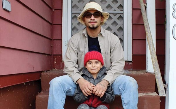 An adult and a young child sit together on brown steps outside a red house. The adult wears sunglasses, a beige hat, and a light jacket; the child wears a gray coat and red knit hat, both smiling at the camera.