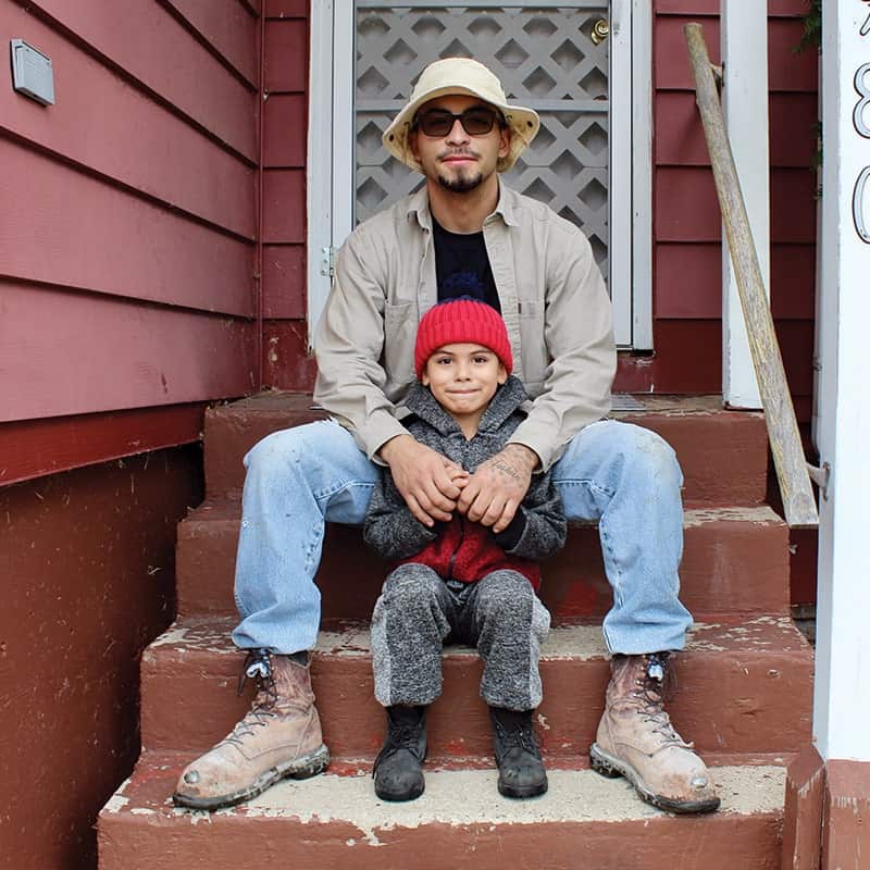 An adult and a young child sit together on brown steps outside a red house. The adult wears sunglasses, a beige hat, and a light jacket; the child wears a gray coat and red knit hat, both smiling at the camera.