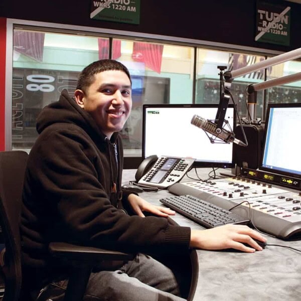 A young man in a hoodie sits at a radio station desk with microphones, computer monitors, and audio equipment, smiling at the camera.