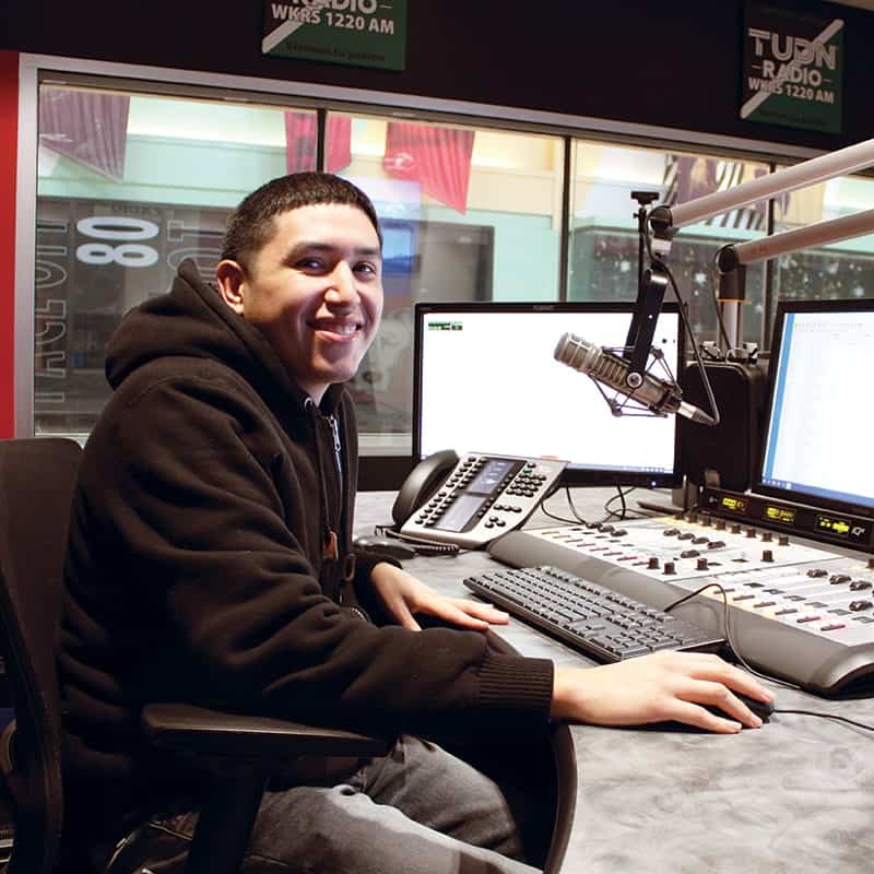 A young man in a hoodie sits at a radio station desk with microphones, computer monitors, and audio equipment, smiling at the camera.